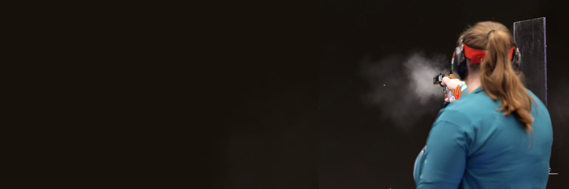 Woman in blue shirt and ear muffs shooting a pardini with muzzle smoke on a black background.