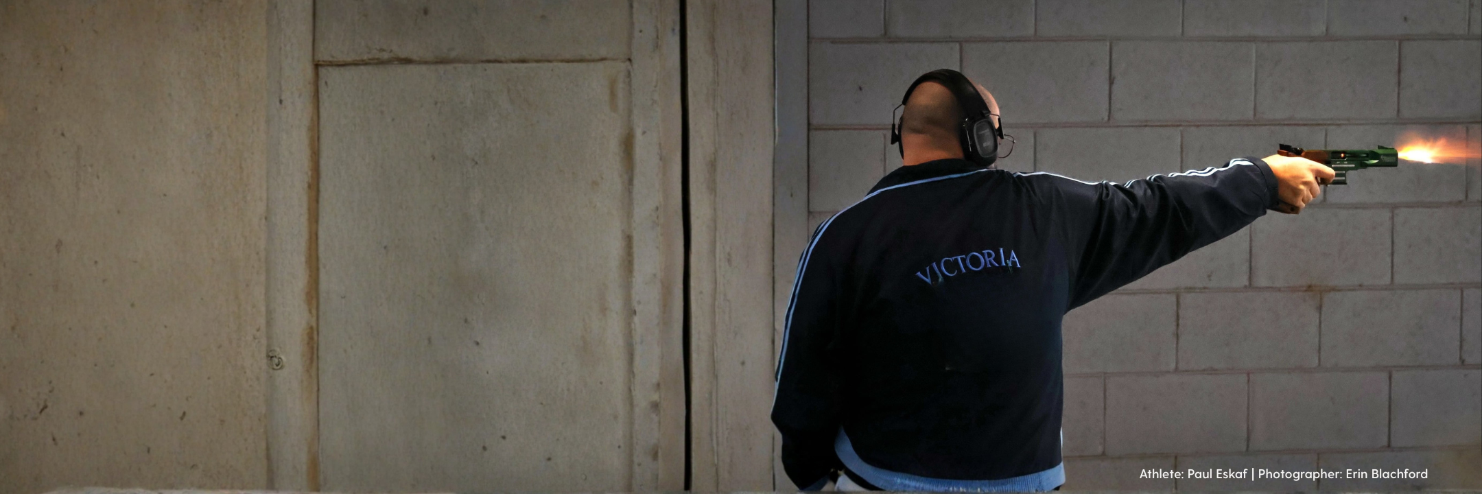 ISSF pistol shooter on indoor range firing shot with visible muzzle flash.